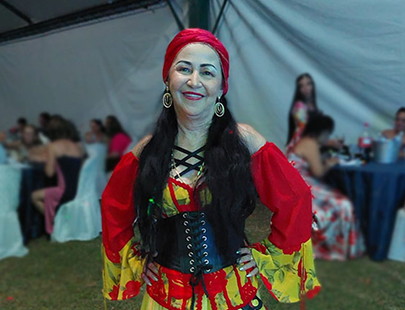 cigana-maura.jpg Foto de uma mulher cigana, sorridente, com vestido e chapeu vermelhos, em uma tenda cigana com de outras ciganas ao fundo