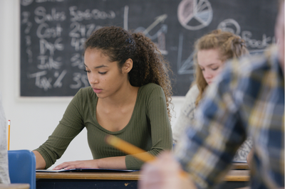 Foto ilustrativa: Canva Foto mostra mulher negra sentada em uma sala de aula, sentada à frente de uma mesa, com os braços apoiados nela. Ela segura um lápis sobre um papel na mesa. Ao fundo e em primeiro plano, desfocados, há uma mulher e um homem branco nas mesma situação.