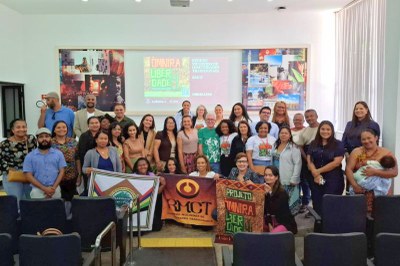 Em uma sala de auditório, um grupo diverso de cerca de 40 pessoas posa sorridente para a foto, de pé e sentadas nas primeiras fileiras. À frente, três faixas são exibidas com os nomes das organizações participantes, incluindo o projeto ÓMNIRA Liberdade e o RMCT – Rede de Mulheres de Comunidades Tradicionais. Ao fundo, um telão exibe o título “ÓMNIRA Liberdade” e informações sobre o evento. As pessoas demonstram alegria e união, em um ambiente iluminado com luz natural que entra por uma cortina lateral.