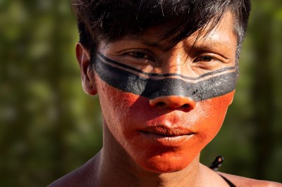 Jovem indígena com o rosto pintado de vermelho e preto, olhando para a câmera.