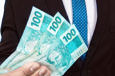 Um homem com traje social, vestindo um terno escuro e gravata azul, segura na mão um leque com três notas de 100 reais brasileiros.