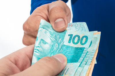 Foto: Canva Foto mostra o detalhe da mão de uma pessoa vestindo terno azul e a mão de outra pessoa (que não aparece na foto) segurando um maço de dinheiro com notas de 100 e 50 reais