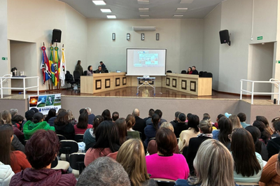 Foto mostra auditório em que a área da plateia tem praticamente todas as cadeiras ocupadas por homens e mulheres e, no palco, à frente do auditório, há pessoas sentadas por trás de bancadas. No centro do palco, há uma tela de projeção