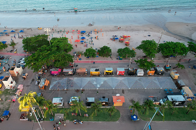 Foto: Polícia Institucional MPF/AL Imagem aérea da orla marítima de Maceió em dia ensolarado. À esquerda, há uma faixa de areia clara com banhistas espalhados e algumas barracas com guarda-sóis coloridos. O mar apresenta tons de azul e verde, com pequenas embarcações ao largo. À direita da praia, há uma calçada arborizada e uma via pavimentada, onde se observa uma fila de food trucks estacionados em paralelo. Esses veículos são utilizados para venda de alimentos e estão posicionados lado a lado, com toldos abertos e pessoas ao redor. O cenário retrata uma área turística ativa, com ocupação informal do espaço público e elementos urbanos contrastando com o ambiente natural da orla.