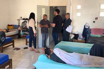 No abrigo Camar 1, em Belém, quatro pessoas conversam em pé. Ao redor delas, há várias camas simples, e em algumas, pessoas estão deitadas descansando.