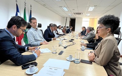 Foto: Comunicação MPF Reunião institucional realizada em sala ampla e iluminada, com 13 pessoas sentadas ao redor de uma mesa retangular de madeira clara. À esquerda, estão representantes do Ministério Público Federal e da Defensoria Pública da União, com papéis à frente e bandeiras da Bahia, do Brasil e do MPF ao fundo. À direita, outros participantes ouvem atentamente. A mesa está organizada com copos d’água, xícaras de café e microfones de conferência. No primeiro plano, sobre a mesa, aparece um documento com os logotipos do MPF e da DPU.
