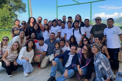Foto: MPF A imagem mostra um grupo diverso de pessoas reunidas ao ar livre. Ao fundo, há uma cerca de tela verde com paisagem montanhosa visível. Algumas pessoas usam camisetas brancas com logotipo escolar.