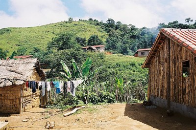 A imagem mostra uma paisagem rural com casas feitas de madeira, barro e telhas. Em primeiro plano, há roupas estendidas em um varal. Ao fundo, vê-se uma área verde com vegetação densa e morros. 