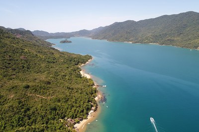 A imagem mostra uma vista aérea de uma área costeira com mar calmo de águas azul-esverdeadas, margeado por uma densa vegetação de Mata Atlântica. Há pequenas praias ao longo da costa, algumas construções rústicas e embarcações ancoradas. Ao fundo, destacam-se montanhas cobertas por floresta.