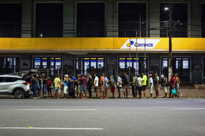 Voluntários distribuem alimentos para pessoas em situação de rua em frente à agência dos Correios, na Av. Presidente Vargas (Foto: Cícero Pedrosa Neto/Amazônia Real) A imagem mostra uma fila de pessoas em pé na calçada durante a noite, em frente a uma agência dos Correios identificada por uma faixa amarela com o logotipo e nome "Correios". À esquerda, um carro prata está estacionado com o porta-malas aberto, onde algumas pessoas se concentram.