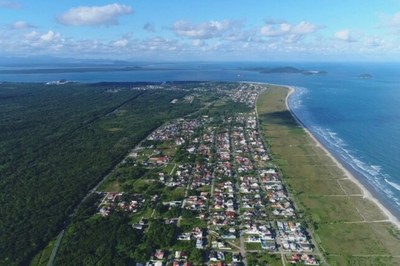 Foto aérea colorida mostra a cidade litorânea Pontal do Paraná, entre o mar e uma grande área de Mata Atlântica. À direita, o mar azul com faixa de areia clara. Ao centro, quarteirões com casas e ruas retas. À esquerda, vegetação densa. No fundo, o mar encontra o céu com algumas ilhas no horizonte.