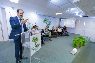 Foto: Leobark Rodrigues/Secom MPF A imagem mostra o procurador da República Wilson Rocha Assis discursando em evento comemorativo dos 25 anos do SNUC, promovido pelo ICMBio. Ele fala ao microfone, em um púlpito com o logotipo do instituto, enquanto cinco pessoas — incluindo um indígena com cocar e servidores do ICMBio — o acompanham sentadas ao fundo. O ambiente é institucional, com decoração simples, plantas e uma tela de projeção exibindo a identidade visual do evento.