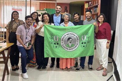 Foto: Comitê Chico Mendes Foto mostra cinco mulheres e quatro homens em pé, lado a lado em ambiente interno, posando para a foto. Uma das mulheres e um dos homens seguram uma bandeira verde à frente, com a imagem de Chico Mendes e a expressão Chico Mendes A luta continua.