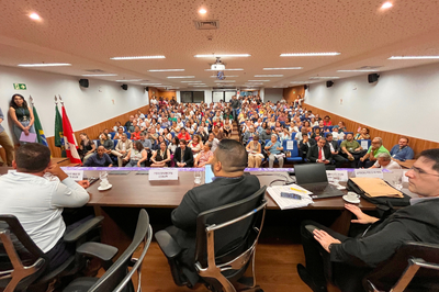A imagem mostra um auditório lotado, com fileiras de poltronas azuis ocupadas por pessoas voltadas para uma mesa no palco. Atrás da mesa estão três homens de costas para a câmera, com laptops, papéis e copos sobre a mesa, e ao centro há uma placa escrita “PROCURADOR DA REPÚBLICA BELÉM”. No canto esquerdo, há três bandeiras, incluindo a do Brasil.