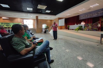 Um homem de camisa listrada em tons de rosa e calça azul fala em pé no centro de um auditório, segurando um microfone e um papel, enquanto três pessoas sentadas em cadeiras pretas na primeira fileira o observam; ao fundo, no palco, há uma mesa coberta com toalha branca, duas pessoas sentadas e um painel colorido com desenho de onça, além de um banner azul com o texto "Audiência Pública " Gestão Social da Bacia do Tapajós e seus Recursos Hídricos " MPPA " MPF".