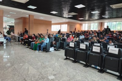 Várias pessoas estão sentadas em cadeiras de um auditório, voltadas para a frente; na fileira mais próxima da câmera há quatro assentos com placas brancas com a palavra ¿RESERVADO¿ em letras maiúsculas; ao fundo, há pessoas operando câmeras e equipamentos, e nas paredes janelas deixam entrar luz natural.