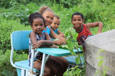 Grupo de três crianças, com uma delas sentada numa cadeira escolar, no meio do mato.