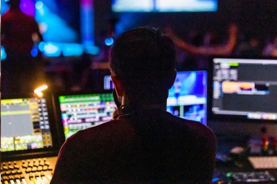 Imagem escura de uma ilha de edição de vídeos mostra diversos computadores com programas de edição abertos nas telas e uma pessoa sentada olhando para tela