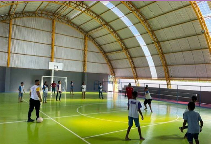 Foto de um ginásio de esportes com teto curvo visto de dentro com crianças brincando na quadra