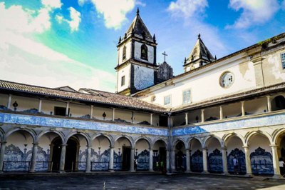 A imagem mostra o pátio interno de um edifício colonial, provavelmente um mosteiro ou igreja, com um céu azul e nuvens brancas ao fundo. No centro da cena, há um pátio pavimentado cercado por uma arcada em dois níveis. As paredes do andar de baixo são decoradas com painéis de azulejos azuis e brancos, um estilo característico da arte portuguesa. No fundo, é possível ver duas torres sineiras brancas, uma delas com uma cruz no topo. A iluminação sugere que a foto foi tirada em um dia claro, e as sombras realçam a arquitetura histórica do local.