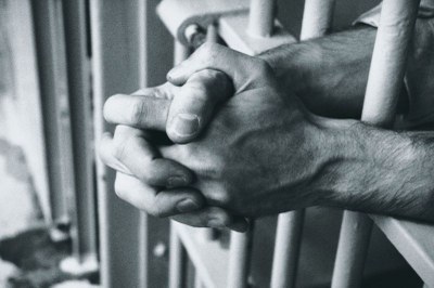 Imagem de uma pessoa dentro de uma cela e as mãos cruzadas 