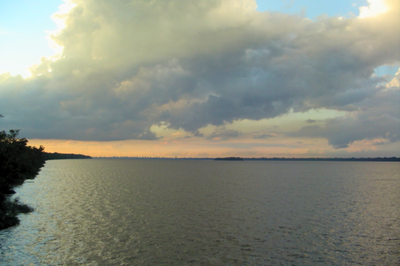 Foto: Gdamasceno Foto mostra um grande corpo de água (a baía do Guajará) com superfície levemente ondulada, ocupando a maior parte do quadro, e uma faixa de vegetação escura à esquerda. O horizonte apresenta uma linha de terra baixa ao fundo e o céu está coberto por nuvens densas e escuras, com áreas iluminadas em tons claros próximos ao pôr do sol.