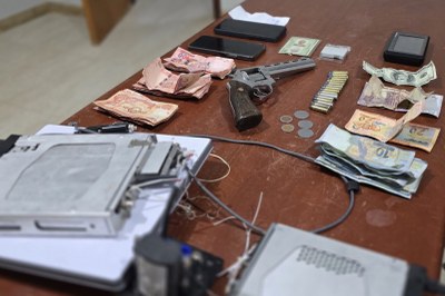 Itens apreendidos com os investigados (foto: PF) Sobre uma mesa de madeira, há um revólver, munições, dinheiro, celulares, documentos e equipamentos eletrônicos dispostos lado a lado.