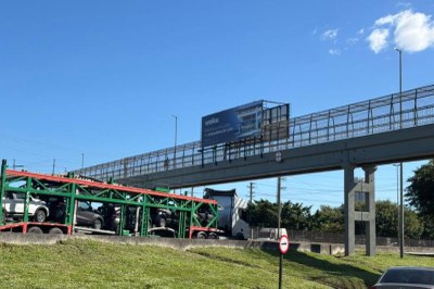 Foto da passarela na BR101 com um painel publicitário e um caminhão cegonheiro passando por baixo 