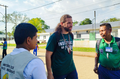 Foto mostra, em área externa, ao centro, um homem branco de cabelos compridos, com uma blusa verde em que se lê "Indígenas no plural" e colares indígenas; ao lado dele, há outros dois homens de pele morena, um deles também usa colares indígenas, o outro usa um colete com a inscrição "Ministério dos Povos Indígenas" nas costas