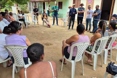 A imagem mostra uma reunião ao ar livre, em um espaço de chão batido, diante de casas simples, pintadas em tons claros de verde e bege. As pessoas estão sentadas em cadeiras plásticas brancas, formando um círculo.

Em primeiro plano, vemos de costas várias mulheres, algumas de cabelo preso e vestindo blusas sem mangas, olhando para o centro da roda. No lado direito, há homens e mulheres em pé, alguns com braços cruzados, chapéus e bonés, acompanhando a conversa.

No centro do círculo, dois homens estão sentados, enquanto outras pessoas em pé parecem conduzir o diálogo. O clima é de escuta e troca de informações, característico de uma reunião comunitária. Ao fundo, árvores e detalhes das casas reforçam o ambiente rural e acolhedor da comunidade.
