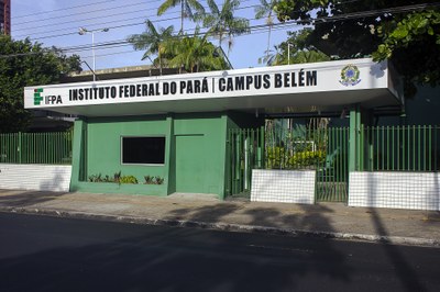 Imagem: Divulgação IFPA A fotografia mostra a fachada do Instituto Federal do Pará, Campus Belém, um prédio térreo verde com uma grande placa branca na entrada. Na placa, está escrito "IFPA INSTITUTO FEDERAL DO PARÁ | CAMPUS BELÉM" em letras pretas, acompanhado do logotipo da instituição e do brasão da República. O local é cercado por uma grade verde sobre uma mureta de azulejos brancos, com árvores e um céu azul visíveis ao fundo. A imagem é capturada a partir da rua, em frente ao portão de entrada.