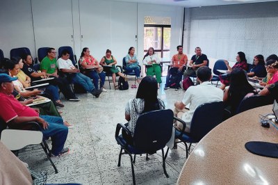 Foto: MPF/AM A imagem mostra uma reunião em andamento, com pessoas sentadas em círculo em uma sala. Os participantes, incluindo mulheres e homens — alguns com vestimentas e acessórios indígenas —, estão atentos, ouvindo ou fazendo anotações. O ambiente é fechado, com piso claro e paredes brancas.
