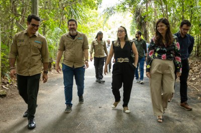 Grupo de pessoas caminha por trilha cercada de vegetação. Alguns usam camisas do Inema, outros estão com roupas casuais. Todos seguem juntos em ambiente de visita técnica ao ar livre.
