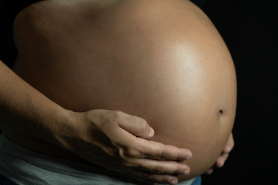 Foto mostra detalhe da barriga de uma mulher negra grávida com as mãos na parte da baixo da barriga; o fundo é escuro