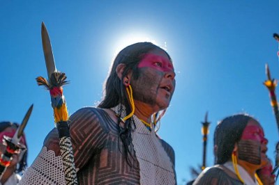 Foto ilustrativa por André Guajajara, via Agência Pará Uma pessoa indígena com o rosto e o corpo pintados em preto e vermelho olha para o lado, em um dia de sol forte. Ela segura uma lança decorada com penas coloridas, e ao fundo, outras pessoas e lanças são visíveis contra um céu azul intenso