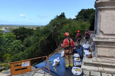 A imagem mostra três bombeiros-militares, equipados com capacetes e cintos de segurança, trabalhando em um terreno inclinado e coberto de vegetação. Eles estão perto do que parece ser a estrutura de um edifício histórico ou uma igreja, indicado por uma parede de pedra ornamentada à direita. O bombeiro mais próximo está segurando uma corda e está em pé sobre sacos de areia e uma lona preta, que aparentemente foram usados para conter algum deslizamento de terra. Outros dois bombeiros estão mais acima na encosta. A paisagem ao fundo é de uma cidade, com prédios e casas, e, mais distante, a orla de uma praia ou lagoa. A luz forte e o céu azul indicam que é um dia ensolarado.