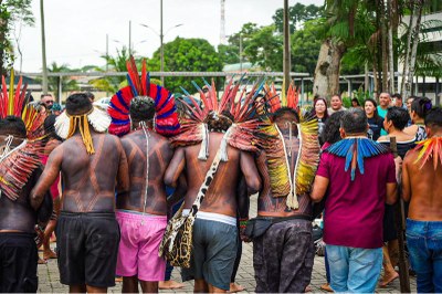 Foto: Jander Arapiun/Cita/Ciki, via Amazônia Real, em licença CC BY-ND 4.0 Foto de grupo de pessoas indígenas de costas, vestindo trajes tradicionais com penas coloridas e pinturas corporais. Elas estão em um ambiente ao ar livre, na área do prédio da Seduc, em Belém.