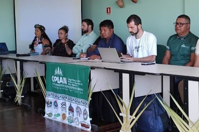 A imagem mostra um painel de pessoas sentadas em uma mesa em um auditório. Há um banner na frente da mesa, que identifica a OPIAM (Organização dos Povos Indígenas do Alto Madeira). A OPIAM participou do evento.