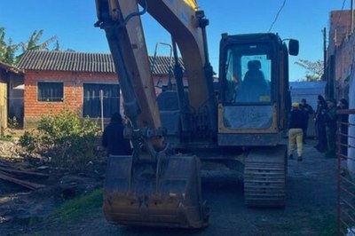 Foto colorida mostra uma escavadeira amarela, utilizada na 24ª fase da Aifa; ao redor da máquina e ao fundo, diversas pessoas, incluindo agentes policiais uniformizados, observam o local.
