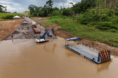 A imagem retrata um trecho de uma rodovia em construção, a BR-364, no Acre, onde a acessibilidade é garantida por uma balsa de metal que faz a travessia de veículos sobre um rio de água barrenta. Na margem oposta, a estrada de terra e cascalho está em obras, com caminhões e uma escavadeira operando. A balsa, com a inscrição "RIO PARANÁ", está ancorada na margem, sugerindo que o transporte fluvial é crucial para conectar as duas partes da rodovia. Ao fundo, a paisagem é composta por densa vegetação e o céu nublado indica tempo instável.