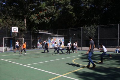 Foto: Rovena Rosa/Agência Brasil A imagem mostra um grupo de jovens em uma quadra poliesportiva ao ar livre, em um dia de sol. Eles estão participando de uma atividade ou jogo com uma bola. Alguns parecem estar na defensiva, enquanto um dos jovens, no centro, prepara-se para rebater a bola com um taco.
A quadra tem o piso verde com linhas amarelas e brancas. No lado esquerdo, é possível ver uma tabela de basquete. A área de lazer é cercada por uma tela de arame e, ao fundo, há árvores, o que sugere um parque ou um ambiente escolar. Há também uma construção de concreto com algumas artes coloridas na parede.
