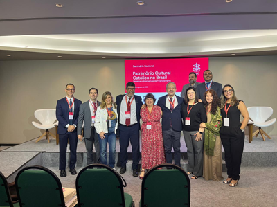 A imagem mostra um grupo de onze pessoas, vestindo roupas de trabalho, posando para uma foto em um palco. O cenário principal é uma tela grande e iluminada, com o texto "Seminário Nacional Patrimônio Cultural Católico no Brasil", acompanhado de uma data e o logo de um evento. Os participantes estão dispostos em duas filas, sorrindo para a câmera. Ao fundo, cadeiras modernas e brancas estão dispostas de cada lado do palco. O ambiente parece ser o auditório de um seminário ou conferência.