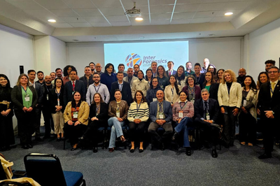 Grande grupo de pessoas, de diversas idades e gêneros, posa para uma foto em um auditório. A maioria veste trajes de trabalho, e ao fundo, um telão com o logo do evento exibe a inscrição "Inter Forensics".
