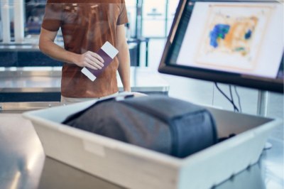 Imagem mostra bagagem passando em equipamento de raio-x em aeroporto. Ao fundo, uma pessoa segura um passaporte na mão