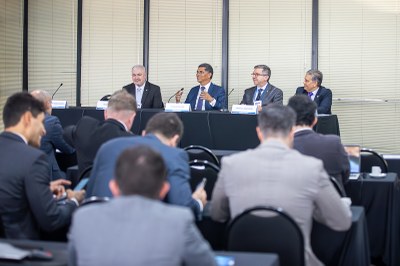 A imagem mostra uma mesa de debate com cinco homens, presumivelmente autoridades, sentados atrás de uma longa mesa preta, voltados para uma plateia. Eles estão vestidos com ternos escuros e camisas sociais. À frente de cada um, há um microfone e um crachá, embora os nomes não sejam claramente legíveis.

A plateia, composta majoritariamente por homens também em trajes sociais, está de costas para o observador, atenta ao que é dito na mesa. O ambiente é de uma sala de conferências, com paredes claras e um banner cinza ao fundo. A iluminação é adequada, criando um clima profissional e sério para o evento.