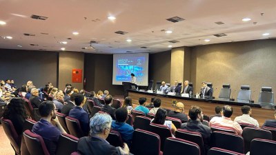 A imagem mostra a palestra Investigação Criminal na Era da Informação. O evento ocorreu no auditório da Procuradoria da República em Teresina (PI), no dia 1º de agosto de 2025. No centro da sala, há uma tela de projeção. Uma pessoa está de pé, próxima à tela. Em frente à tela, há uma mesa comprida onde estão sentadas cinco pessoas, todas homens, com vestimentas sociais (ternos). O auditório está ocupado por uma plateia que parece ser formada por profissionais e estudantes. A maioria está sentada de costas para a câmera, prestando atenção à apresentação. As cadeiras são estofadas, em tons de vermelha.
