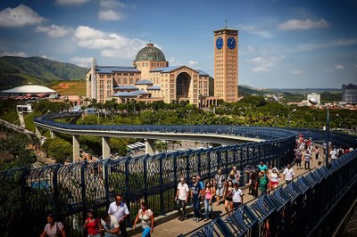 Foto ilustrativa: Ken Chu/Secretaria de Turismo de São Paulo A imagem mostra a Basílica de Nossa Senhora Aparecida, em Aparecida (SP), ao fundo, com sua arquitetura em tijolos aparentes, cúpula central e torre com relógio azul. Em primeiro plano, há uma passarela elevada repleta de fiéis e visitantes caminhando em direção ao santuário. Ao redor, há vegetação, construções e colinas sob um céu azul com poucas nuvens.