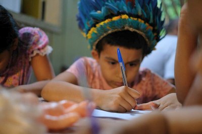 Foto de garoto indígena que usa cocar de penas azuis e amarelas e está concentrado enquanto escreve com uma caneta em um papel sobre uma mesa.
