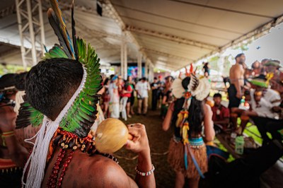 A imagem mostra uma celebração indígena sob uma grande tenda, com pessoas em trajes tradicionais e cocares coloridos. Em destaque, um homem segura um maracá, com penas verdes ao redor da cabeça. Ao fundo, há uma multidão acompanhando o evento