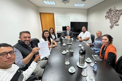 A imagem mostra um grupo de oito pessoas, aparentando ser de diferentes idades e gêneros, sentadas em uma sala de reuniões. Eles estão ao redor de uma grande mesa preta, onde há xícaras de café, um bule, copos e garrafas. Uma pessoa, no lado esquerdo e mais próxima da câmera, segura e mostra um documento. O grupo parece estar engajado em uma conversa ou reunião, com algumas pessoas sorrindo para a câmera. Ao fundo, há uma parede com um grande mapa do Brasil em relevo, e uma porta marrom.